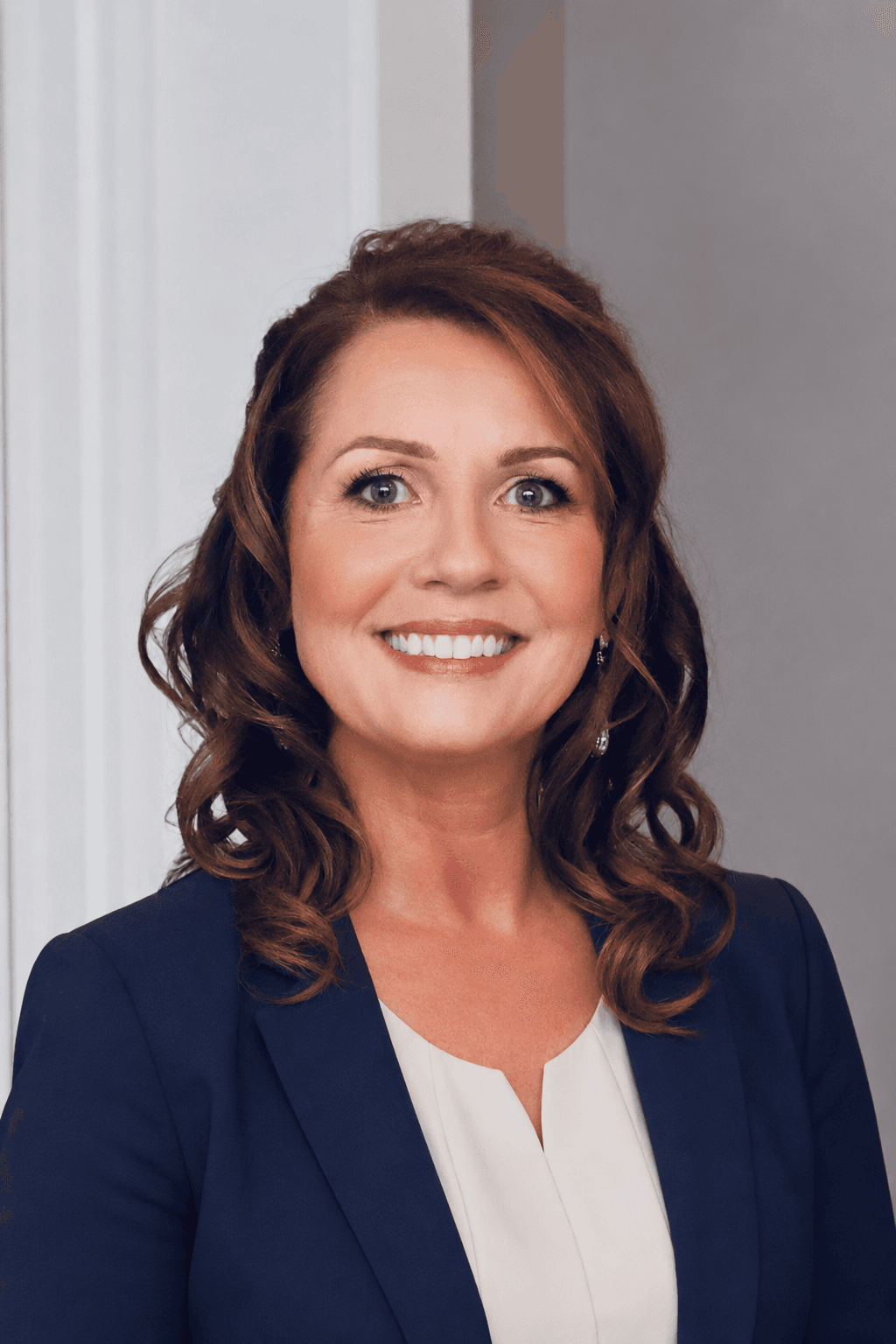 Smiling woman with wavy brown hair wearing a navy blue blazer and white blouse.