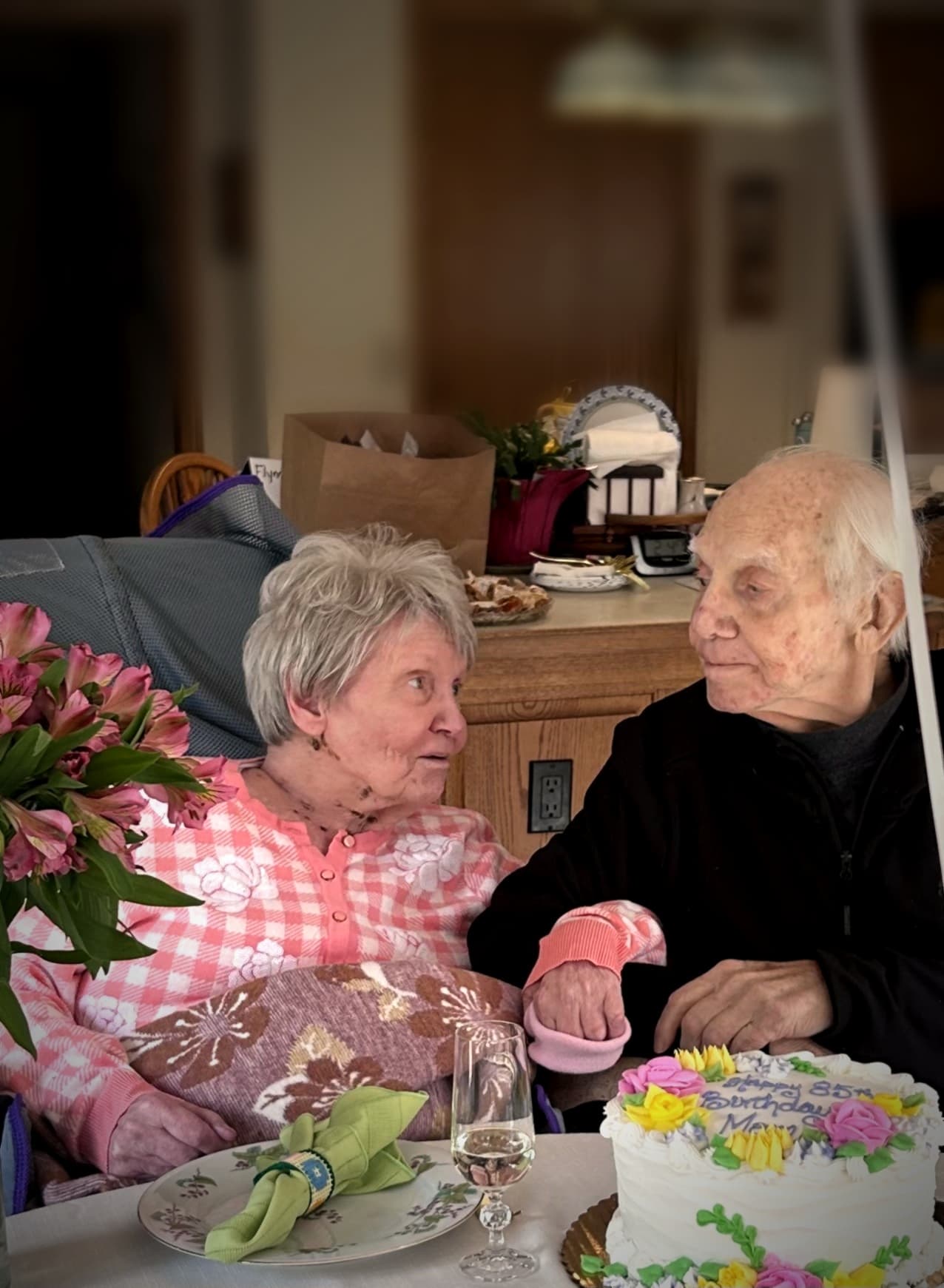 Elderly couple looking at each other behind an 85th birthday cake and pink flowers.